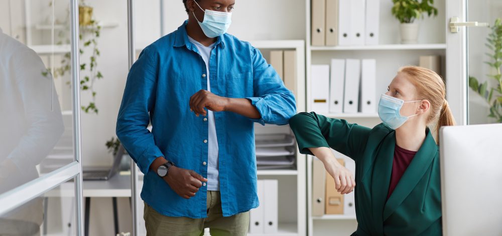 Portrait of young African-American man wearing mask bumping elbows with female colleague as contactless greeting in post pandemic office, copy space