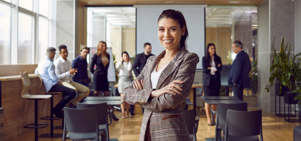 Happy professional business teacher after corporate training class for team of workers. Beautiful young woman in suit jacket standing in office conference room, looking at camera and smiling