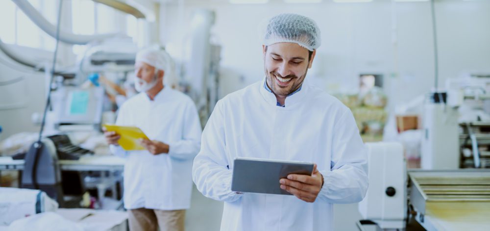 Young Caucasian smiling supervisor in sterile white uniform using tablet while standing in food plant. In background older one worker controlling machine.